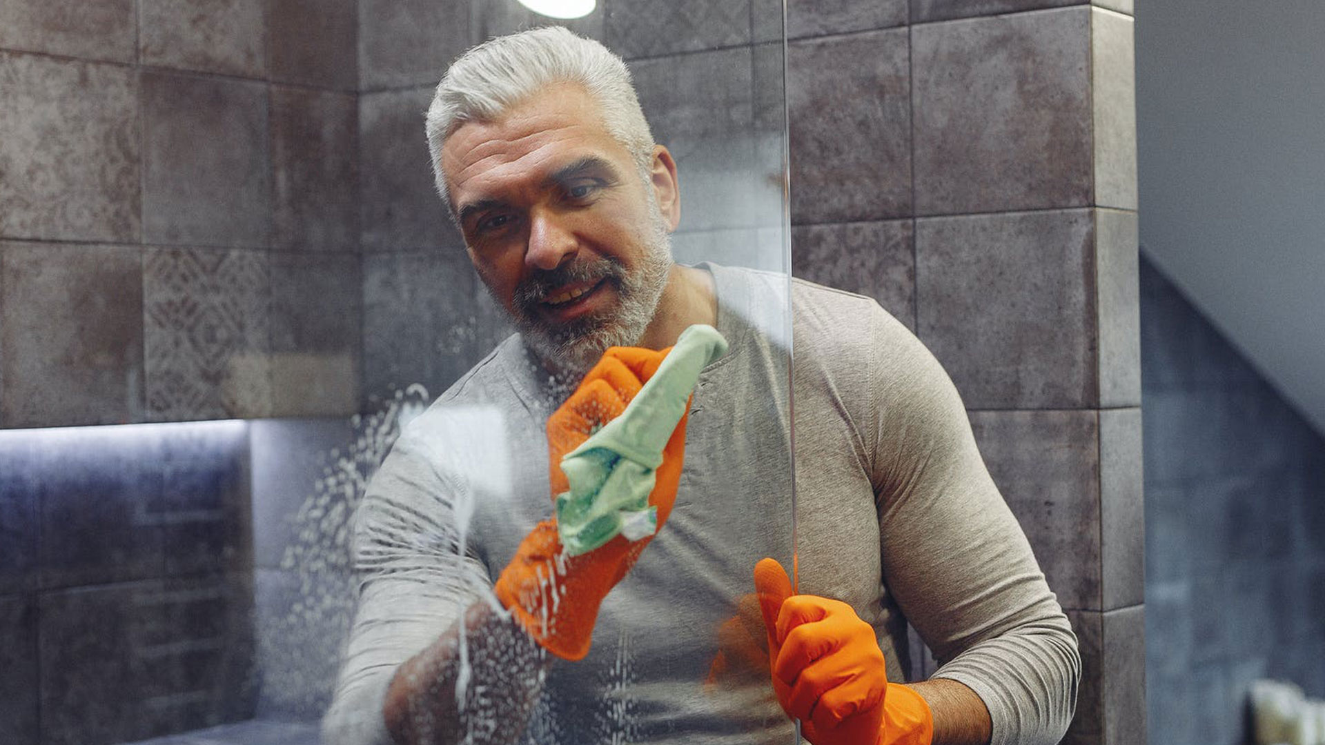 Un homme qui lave ses vitres de salles de bain avec du produits et une serviette. Il porte des gants orange,, un haut gros et il a les cheveux blancs. 