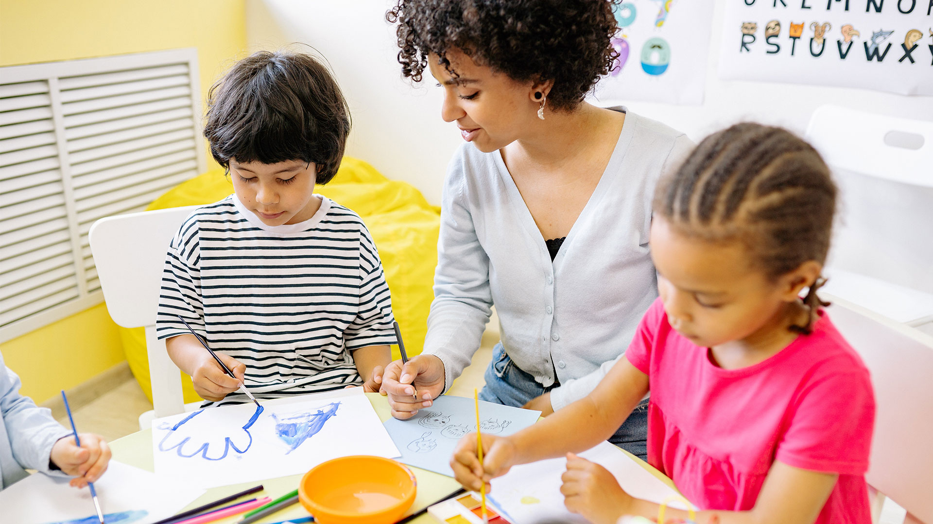 Une image représentant des enfants avec une dame qui s'occupent d'eux. Les enfants dessinent avec l'aide de cette dame.