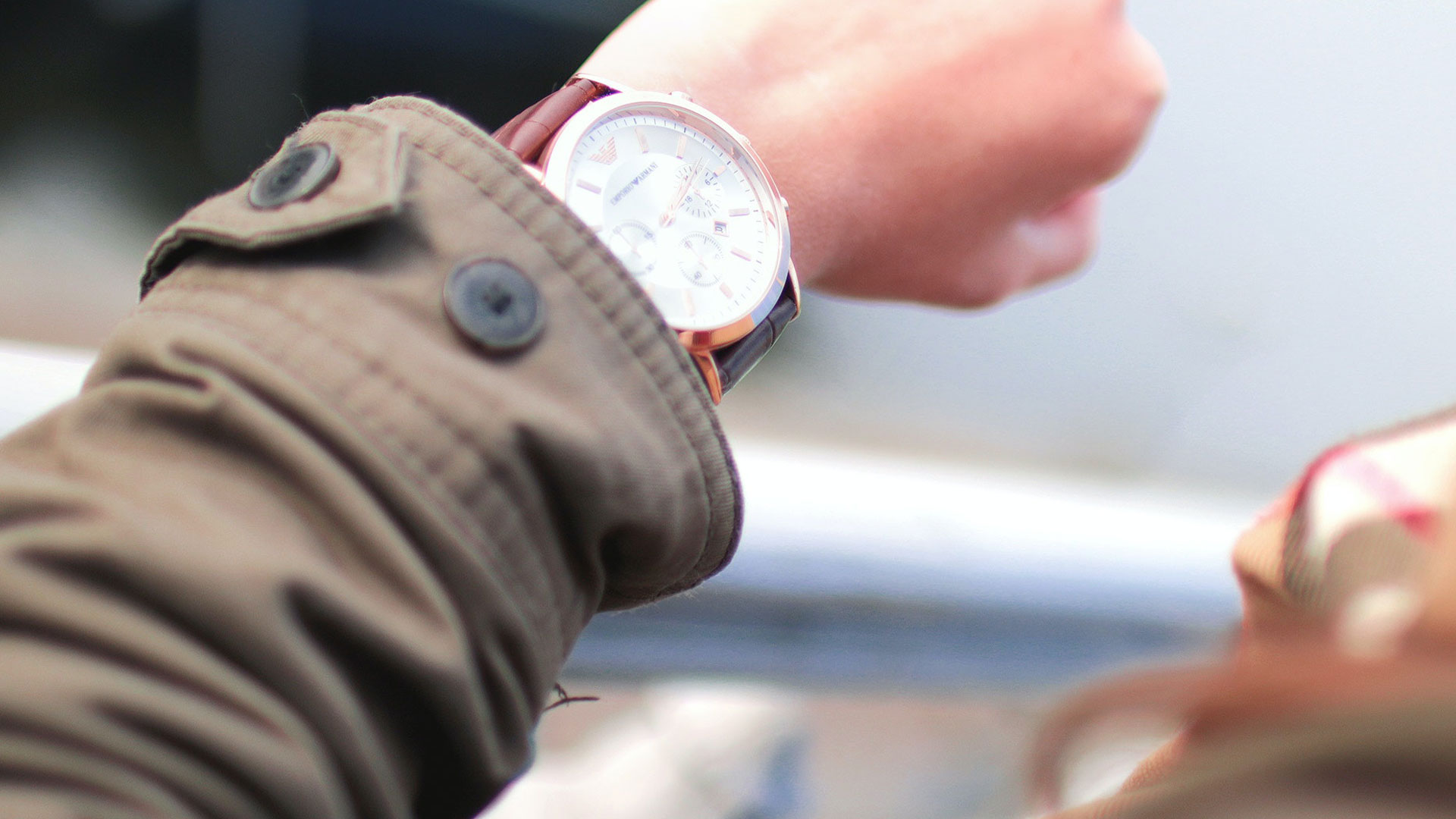 La photo représente une personne qui regarde sa montre. La veste de la personne est kaki et la montre est marron. 