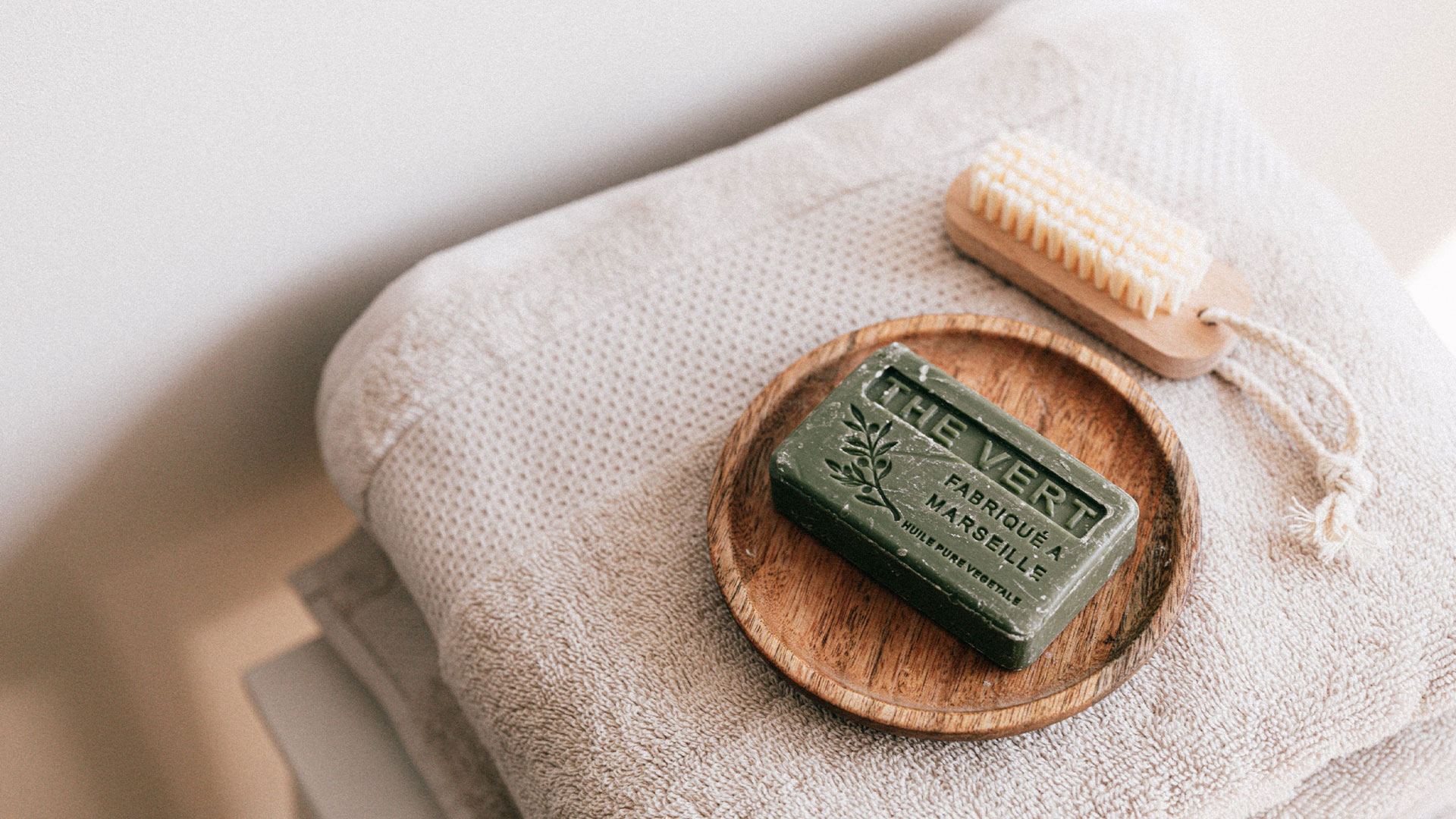 L'image représente un savon vert de Marseille dans un contenu en bois. Ce contenu en vois est sur des serviette de bain avec une brosse à sa droite. La photo est très épurée. 
